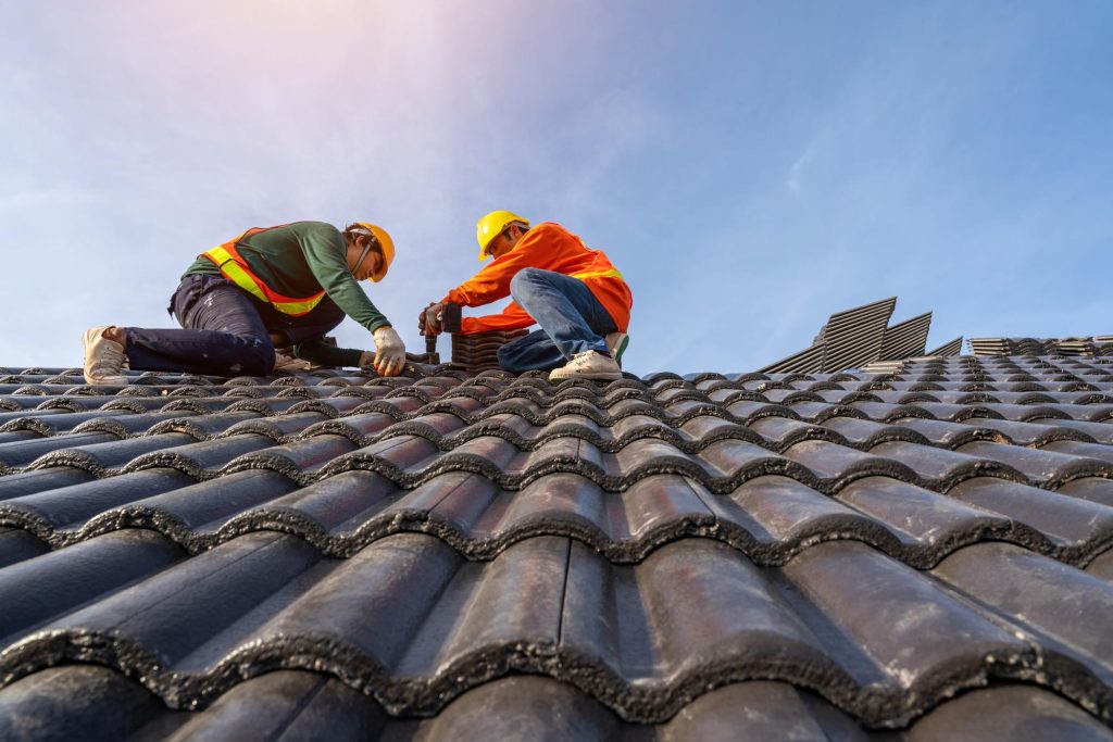 A team of Asian roofing professionals in protective uniforms and gloves, using an air pneumatic nail gun to securely install concrete roof tiles on the top of a new building under construction
